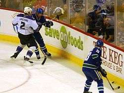 Two ice hockey teammates in the process of passing to one another, while an opposing player checks one of them into the boards. The teammates are dressed in a blue jersey, while the opposing player is dressed in white.