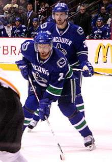 Two twin ice hockey players in ready positions on the ice, lining up for a faceoff. One player in the forefront, is leaning forwards, putting his weight on his stick on the ice, while the other player in the background is standing upright. They are both looking intently forward. They wear blue jerseys with white trim and blue visored helmets.