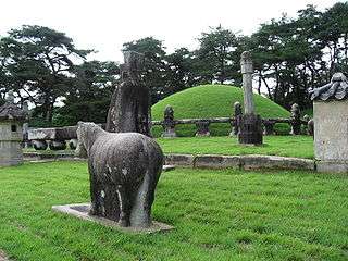 A stone sculpture of a horse and a man, a stone column and a grass covered mound surrounded by a carved stone fence.