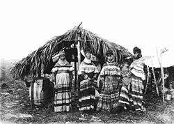 A black and white photograph of four Seminole women and a child standing in front of a chickee wearing bright cotton Seminole patterns