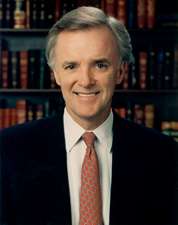 A color image of Kerrey from the waist up wearing a suit and tie. He is smiling at the camera and there is a book shelf with books behind him.