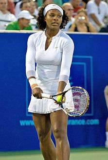 An African American woman in all white clothing, shirt, bandanna, and skirt, is starting the serve of a tennis ball