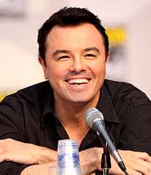 A man with short black hair and a black shirt in front of a microphone. His arms are crossed, and he is laughing.