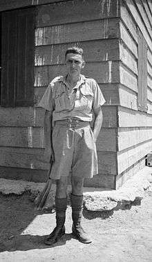 A man, John Hinton, dressed in tropical/desert military uniform and standing outside a hut