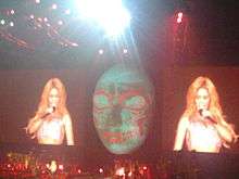 A large green coloured face with red markings is being projected onto a large screen. On both the sides of the face, a woman with long blonde hair is singing with a mic in her hand.