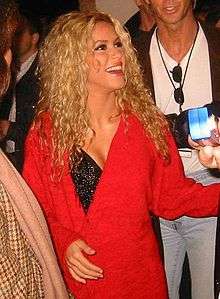 A woman with curly golden hair is dressed in a red jacket over a black top. She is looking upwards and laughing.