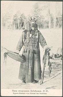 Faded black and white photo of a Mongol Buryat shaman, a masked man wearing a thick robe, an apron covering his chest, and a round hat with covering his face down to his nose. He is holding a drum in his right hand, and two decorated wooden sticks in his left hand. The caption in Russian and French means "Buryat shaman."