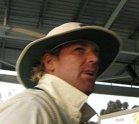A man in cricket whites and hat. He is standing on steps of a stand.