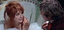 A color screenshot from the film, The Fearless Vampire Killers. Tate is sitting in a large ceramic bathtub, filled with bubbles up to her shoulders. Strands of hair from her red wig are draped over her face, as she looks, smiling, at Roman Polanski, who is leaning towards her at the side of the bathtub.
