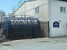 A large, cylindrical tank with the words Doom Bar written on it, lying horizontal behind the fence of an industrial unit