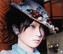A woman wearing a straw hat with ornamental fruit and blue contact lenses stares off to her left.