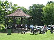 Grassy area with people sitting in chairs in from of a bandstand with tiled roof.
