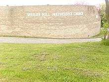 Behind a large area of mown grass stands a beige brick wall which curves around a corner to the right. The wall bears the words "Sheriff Hill Methodist Church", which follow the curve of the wall.