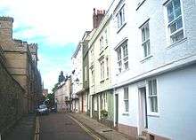 A narrow road, pavements either side; on the left, a stone wall; on the right, a row of terraced three-storey houses, the upper storeys overhanging the ground floor