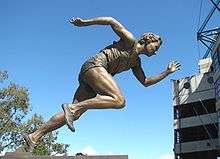 bronze statue of a woman moving from a crouched running position into an all out spring.  Statute us on a black pedestal. It is located outside against a clear blue sky.