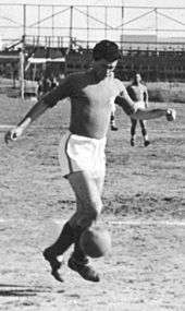 A black-and-white photograph of an athletic-looking association football player of Semitic appearance, wearing a dark shirt, white shorts and dark socks and kicking an old-fashioned football with his right foot