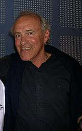 A photograph of a white man with balding grey hair, smiling at the camera while wearing a black buttoned shirt with a blue jacket.