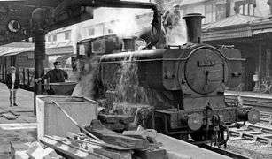 A pannier tank locomotive is standing at a station platform and taking on water from a large filling pipe. The fireman is standing on the locomotive. The driver is controlling the water valve, but may have been distracted by the photographer, as water is cascading over the side of the locomotive. A boy watches.