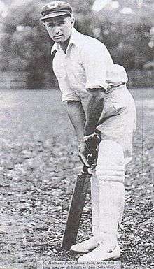 A teenage boy leans over his bat, in front of the stumps, in readiness to face a ball. He is wearing a white shirt, trousers and a cap.