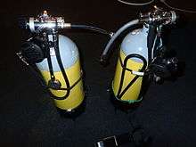 A pair of cylinders showing the regulators set up for sidemount diving. Each regulator has a short low-pressure inflator hose projecting towards where the diver's body would be, and the DV hoses are stowed under bungees. The submersible pressure gauges are on short hoses aligned with the cylinder axes.