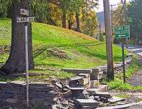 A street corner with the lower portions of a telephone pole and a tree visible above a small stone retaining wall that has crumbled in the center of the photo. On the left is a black and white street sign with "Main Street" on top and "Lake Street" on bottom. At the right a white on green sign has the words "Town of Halcott" and an arrow pointing to the left