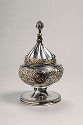 Silver gilt vessel with conical lid. It was intended for use as a ciborium. The vessel is decorated with foliate scrolls and medallions. The knob on the vessel's lid is in the shape of a bunch of grapes.