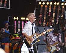 Gillies at left behind his drum kit. Johns is singing wide-mouthed into a microphone and playing his guitar, he wears a garter with a ribbon over his pants on his right thigh. Joannou is playing his bass guitar. In the background are three female singers with one partly obscured at right.