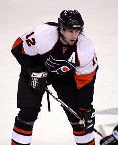 An ice hockey player is leant over slightly. He is wearing a black uniform with white shoulders and orange piping, and '12' is visible on his sleeve. A stylised 'P' is on his chest, and just above it, a smaller 'A'. He is wearing a black helmet with a visor.