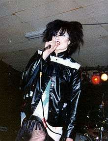 A black-haired woman wearing a leather jacket sings into a microphone.