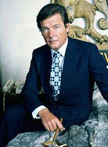 Roger Moore sits facing the camera in a relaxed pose, a cigar held in his right hand.