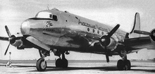 Close-up, in black-and-white, of a silver-bodied four-engined propellor aircraft standing on an apron at an airport