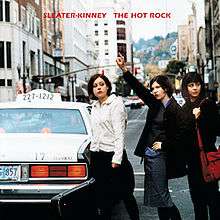 Three women standing on a road. Text above the image reads "Sleater-Kinney The Hot Rock".