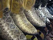 Five pairs of cowboy boots in a row facing half-right, all having snake-skin uppers. The first three pairs have uppers of gray and tan scales and ankle pieces of ecru colour. The last two pairs have uppers with prominent black and white diamond shaped scale patterns and navy blue ankles.