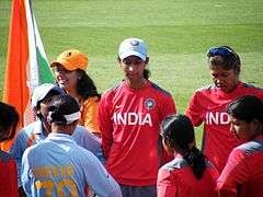 Eight female cricketers stand on a field. Two players are in red practice jerseys; three other players in red jerseys are facing toward them; a player in a blue shirt is facing away and hides another player in a blue game shirt. In the upper left corner is a fan with an Indian flag.