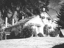 A modest two-story farmhouse with a peaked roof and four chimneys sits under a large tree at the top of a sloping lawn. Bushes and shrubs grow near the house on its two visible sides. A small part of a neighboring house, perhaps only 15 feet (4.6 m) away, can be seen in the background.