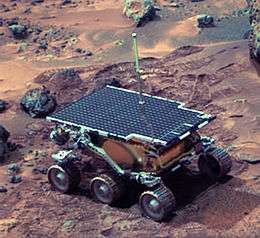 A Mars rover, topped with solar panels, on reddish-brown terrain.