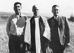 Three men stand in a field. The left-most is casually dressed and of aboriginal heritage, the middle one is a white man wearing ecclesiastical robes, and the right-most is a white man wearing a three piece suit.