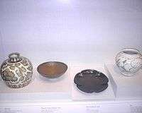 From left to right, a white, nearly spherical jar with a small top and a brown floral pattern glazed onto it, a shallow, unadorned, brown bowl, an unadorned black plate in the shape of a five petaled flower, and a white jar with a large opening and a grey vine pattern glazed into it.
