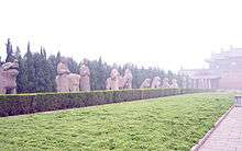 A number of stone statues, mostly of humans and lions, flanking a long paved road that leads to a large building obscured by fog or haze. The statues are separated from the road by a short hedge.