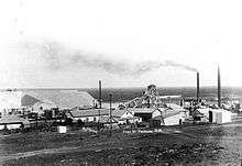 A black and white photograph of a busy mine with several smoking chimneys and industrial buildings