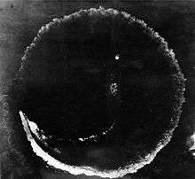Photograph looking directly down at the ocean's surface from a high altitude. There is a rough circle on the surface that is about as wide as the photograph. The top deck of a ship is seen at the lower left of the circle; the length of the ship is much smaller than the circumference of the circle.