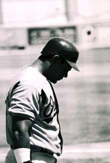 A black-and-white photo of a dark-skinned man in a baseball uniform. His face is partially obscured by the shadow of his batting helmet. His white uniform reads "Orioles" across the chest (obscured).