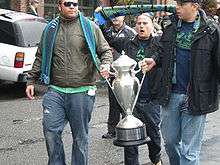 Two men carry a bright silver trophy with another man walking behind holding a green and blue scarf overhead.