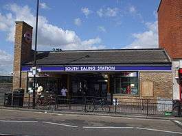 A brown-bricked building with a rectangular, dark blue sign reading "SOUTH EALING STATION" in white letters all under a light blue sky
