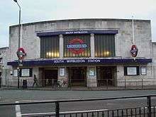 A wide two-storey stone-faced building has three square entrances at the centre beneath a dark blue awning with the words "SOUTH WIMBLEDON STATION". Above the awning is a wide glazed screen in three panels, the centre one of which contains the Underground roundel of a red ring with a blue bar and the word "UNDERGROUND". Two smaller roundels either side of the entrances are mounted on poles at right angles to the face of the building.
