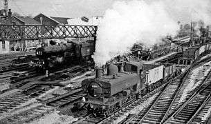 Two steam engines are passing through a station. On the left is a large passenger express locomotive, and on the right, slightly ahead, is a pannier tank locomotive pulling a goods train of open wagons and a flat bogey. It looks as though the locomotives are racing, but as the plumes of steam from the locomotives are at different angles, the fast express will probably quickly catch up with the pannier and pass it.