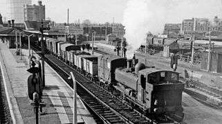 A pannier tank locomotive, travelling in reverse, is pulling a train of open and closed goods wagons through a station.