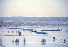 Photograph from elevation of flooded river flowing between snow covered fields. Hills in the distance.