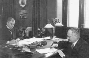 Two men sign papers at opposite sides of a table in a small office.