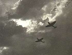 Two Spitfire single-seat piston-engined fighters in flight,  silhouetted against cloud